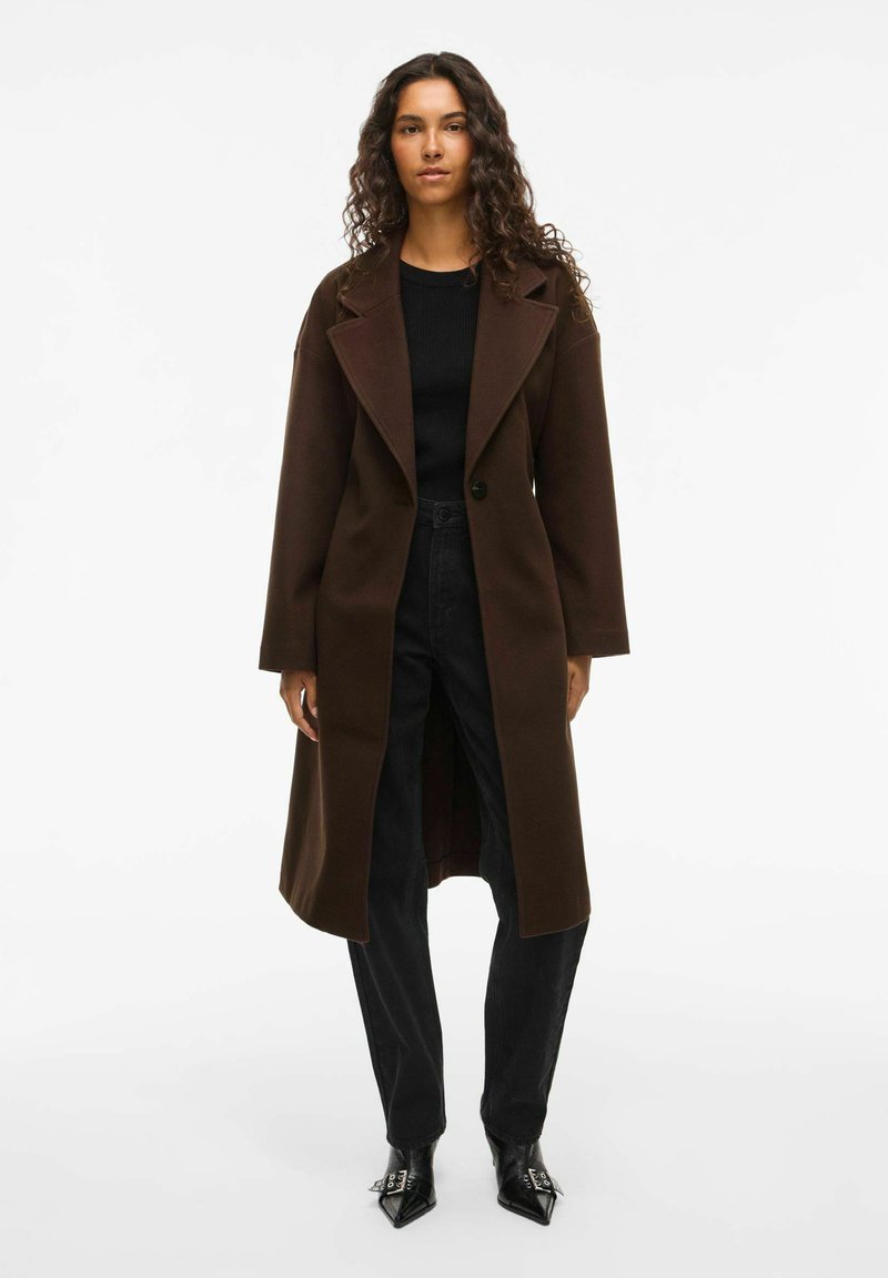 Manteau en mélange de laine marron avec un col cranté, fermeture à un bouton, fentes sur les côtés et coupe décontractée, associé à un pantalon noir texturé.