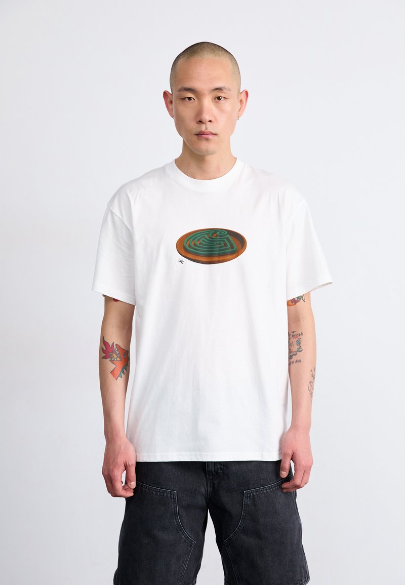 Hvit bomulls t-skjorte med et sirkulært grafisk design av en grønn labyrint med brune aksenter, korte ermer og avslappet passform. Mørke shorts brukt sammen med den.