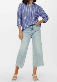 Camicia azzurra a righe con maniche arrotolate, abbinata a jeans in denim a gamba larga di lavaggio chiaro e sandali bianchi. Tessuti texturizzati, design casual.