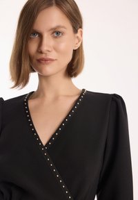 Czarna sukienka o kroju kopertowym z głębokim dekoltem w kształcie V, zdobiona złotymi ćwiekami wzdłuż krawędzi, z bufkami na długich rękawach, wykonana z gładkiego materiału.