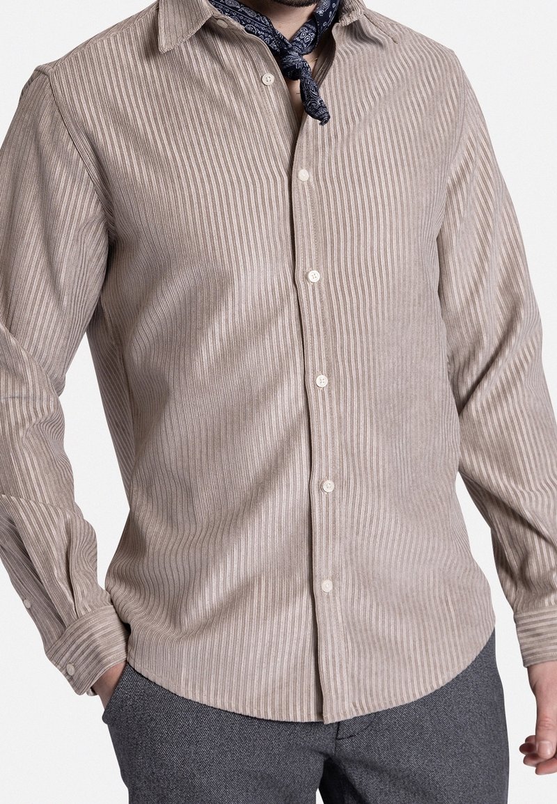 Camisa beige claro abotonada con rayas verticales, mangas largas y cuello clásico. El diseño presenta una textura sutil, complementada con una bufanda oscura.