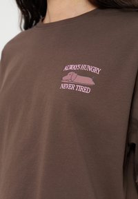 Chemise marron avec un petit graphisme rose d'un chien endormi et un texte au-dessus et au-dessous indiquant "ALWAYS HUNGRY NEVER TIRED" sur la poitrine.