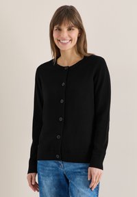 Femme souriante portant un cardigan noir boutonné à manches longues et un jean bleu, debout devant un fond clair uni.
