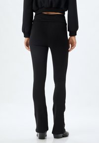 Pantalons noirs évasés avec une texture lisse et une taille haute, présentant un ajustement près du corps au niveau des hanches et un léger évasement à partir du genou.