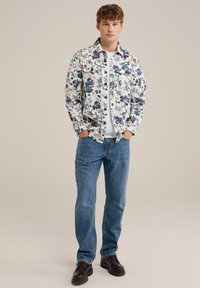 Chemise à boutons à motifs floraux en blanc, bleu et vert ; associée à un t-shirt blanc, un jean bleu et des chaussures noires.
