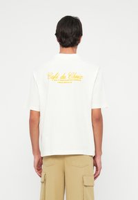 White cotton t-shirt featuring a yellow printed design on the back that reads “Café du Choix 24/7 Breakfast Diner Free Refills.”