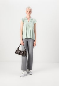 Top corto verde chiaro con maniche corte e scollatura a V, ricamo floreale, abbinato a jeans larghi grigi e sneakers bianche, mentre tiene una piccola borsa scura.