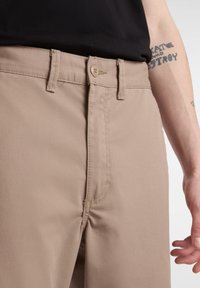 Pantaloni beige in cotone con vestibilità standard. Presentano una chiusura a bottone, passanti per la cintura e dettagli cuciti sulle tasche anteriori.