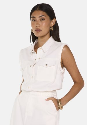 Femme aux cheveux foncés portant un haut blanc sans manches à boutons et un pantalon blanc, accessoirisée avec des boucles d'oreilles créoles dorées et des bracelets.