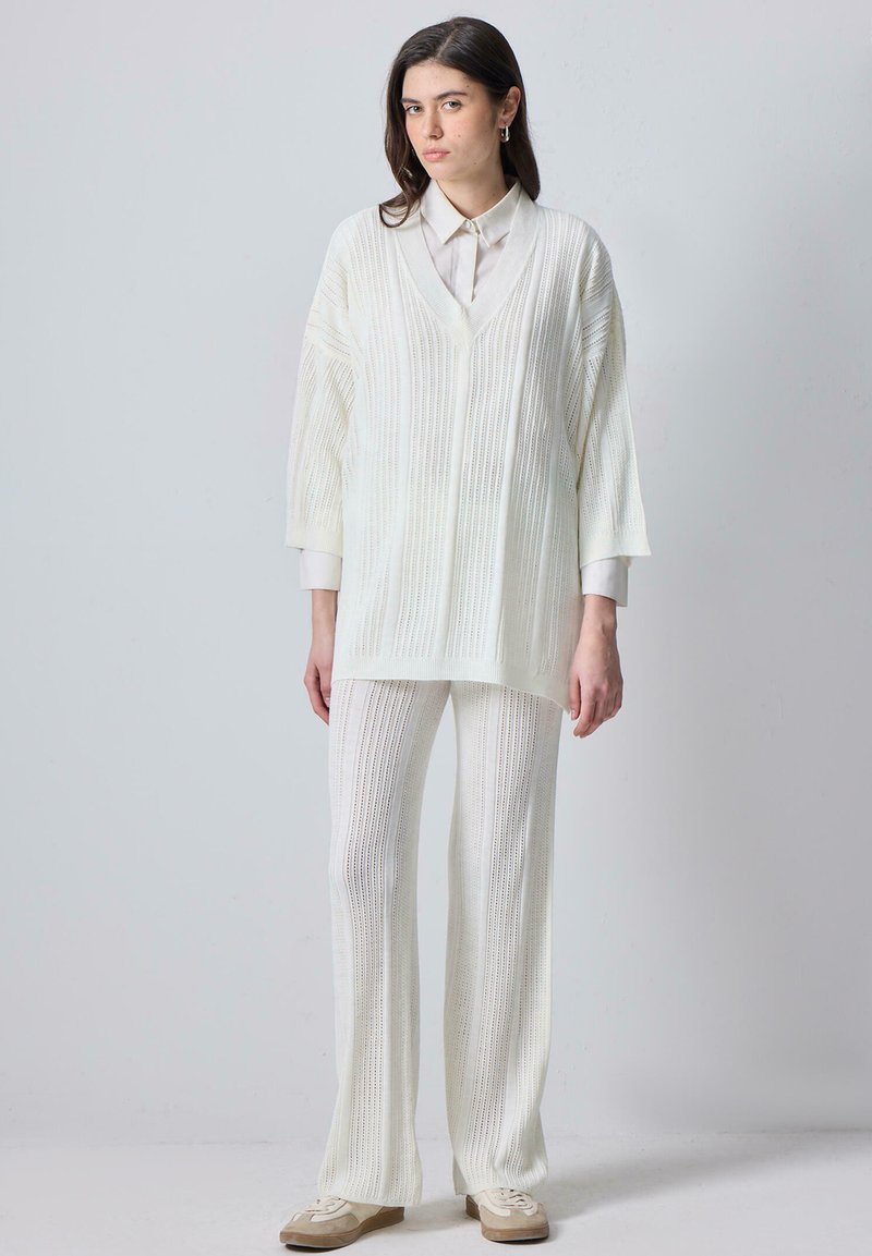 Femme debout portant un pull en maille côtelée blanc, un pantalon large assorti, des baskets beige, et une chemise blanche à col, sur un fond clair uni.