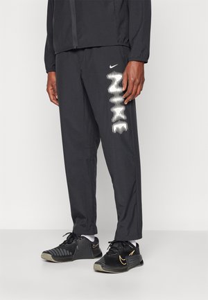 Pantalones deportivos negros con bolsillos laterales, que presentan un patrón geométrico blanco. Combinados con zapatos negros que muestran un logo dorado.