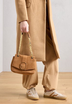 Persoon draagt een beige jas en broek, houdt een bruine suède handtas met gouden ketting vast, staat op een houten vloer in witte sneakers.