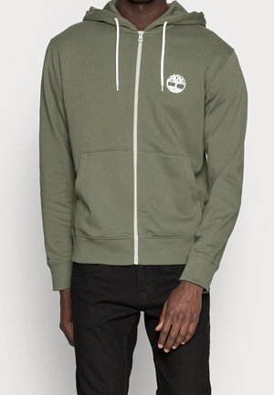 Sudadera con cremallera de color verde olivo, con cordones blancos y logo, que cuenta con un bolsillo tipo canguro y puños y dobladillo acanalados. Textura de tela suave.