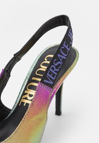 Escarpin slingback à talon haut avec finition irisée, bride noire avec inscription "VERSACE JEANS COUTURE" en violet et marquage doré à l'intérieur.