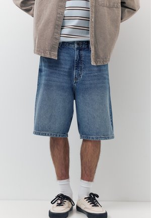 Mann trägt kniehohe blaue Jeansshorts, weiße Socken, cremefarbene Sneakers mit schwarzen Schnürsenkeln, gestreiftes Hemd und beige Jacke.