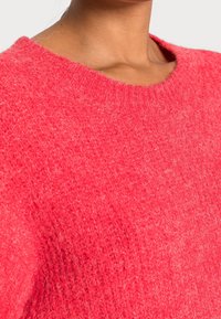 Rosa stickad tröja med rund halsringning, med ett texturerat, lurvigt material och en avslappnad passform. Subtila färgvariationer ger djup.