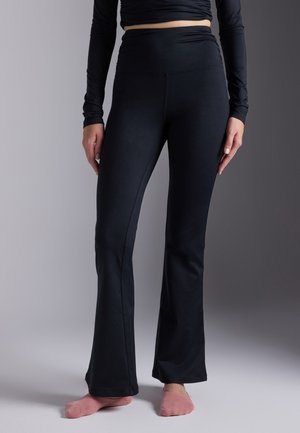 Vrouw draagt zwarte hoog getailleerde uitlopende broek en een zwart cropped top met lange mouwen, staat op blote voeten op een grijze ondergrond.