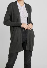 Dunkelgrauer Cardigan aus weichem Material, offene Vorderseite, lange Ärmel, mit gerippten Bündchen und reicht bis zur Mitte des Oberschenkels. Getragen über einem gemusterten Oberteil.