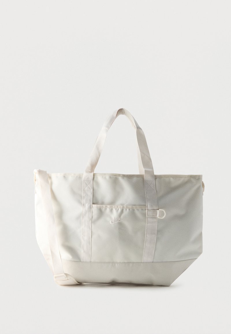 Weiße Nike Tote-Tasche mit zwei Griffen, einer Vorderseite-Tasche und einem verstellbaren Schultergurt vor einem schlichten Hintergrund.