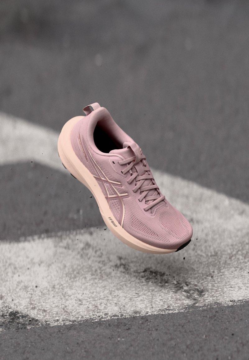 Zapatilla de correr rosa claro en el aire sobre asfalto con marcas de carretera blancas, mostrando una malla transpirable y una suela acolchada con tecnología FP Blast.