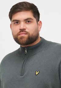 Pull gris zippé avec col côtelé, comportant un petit logo jaune brodé sur la poitrine gauche. Texture en tricot douce, vue de face complète.