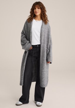 Grijze lange cardigan van een zachte brei met oversized mouwen, voorzakken en geribbelde manchetten, gecombineerd met een witte top en zwarte wijde broek.