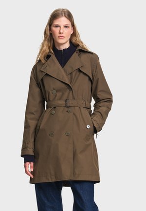 Femme portant un trench-coat ceinturé vert olive par-dessus un pull sombre et un jean bleu, debout avec une main dans la poche devant un fond uni.