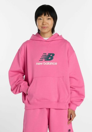 Junge Person trägt ein knallpinke New Balance Hoodie und passende Hose und steht mit einer Hand in der Tasche vor weißem Hintergrund.