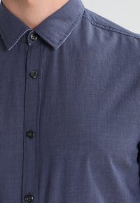 Mężczyzna w niebieskiej koszuli w kratę z guzikami i kołnierzykiem, odsłaniający górną część klatki piersiowej i szyję.