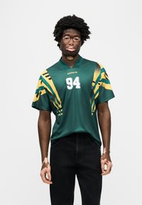 Πράσινο αθλητικό πουκάμισο από πολυεστέρα με κιτρινόμαυρα γεωμετρικά σχέδια, σε φαρδιά γραμμή, με λευκό αριθμό "94" στο μπροστινό μέρος και το λογότυπο της adidas.