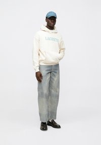 Camisola com capuz creme com "LACOSTE" em azul, boné de ganga azul claro, jeans de perna larga desbotados e sapatos escuros sem atacadores. Aparência limpa e minimalista.