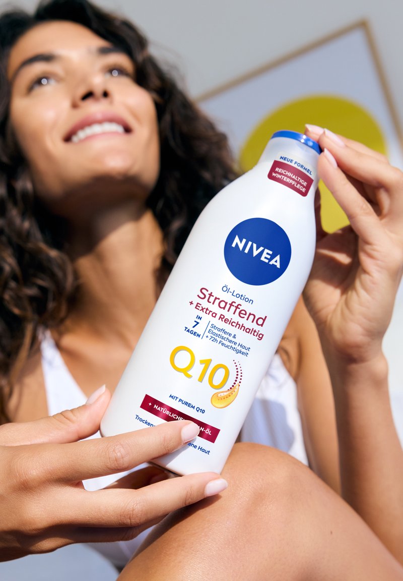 Frau hält eine Flasche Nivea Q10 Öl-Lotion und zeigt die Produktdetails. Sie lächelt mit lockigem Haar und freiem Schulterbereich in hellem Umfeld.