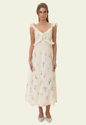 Kobieta ubrana w białą sukienkę midi bez rękawów z falbankowym dekoltem i wielokolorowym kwiatowym wzorem, stojąca z ramionami swobodnie opuszczonymi wzdłuż ciała.