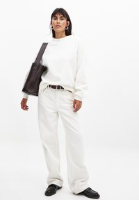 Sudadera blanca de gran tamaño, pantalones anchos color crema y mocasines negros. Un bolso de mano marrón oscuro cuelga de un hombro. Acentos de anillo simple en plata.
