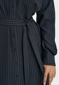 Črna obleka s črtastim vzorcem, z gumbi spredaj, s pasom na vezanje in dolgimi manšetnimi rokavi, prikazana na trup in roki osebe.