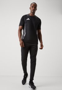 Heren zwart sportoutfit met T-shirt met korte mouwen en broek. Bevat grijze accenten en een logo met drie witte strepen op het shirt. Sportschoenen.