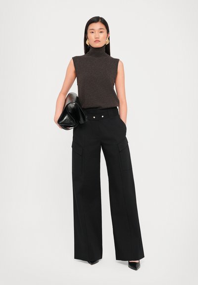 Brown sleeveless turtleneck sweater, high-waisted black wide-leg trousers with front pockets, and black clutch bag. Plain background.