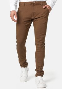 Brune tætsiddende bukser lavet af struktureret stof, med knaplukning, sidelommer og parret med hvide sneakers.