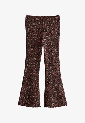 Ung pige med langt hår iført en sort top med lange ærmer, brune leopardmønstrede bukser og sorte og hvide sneakers, der står mod en ensfarvet baggrund.