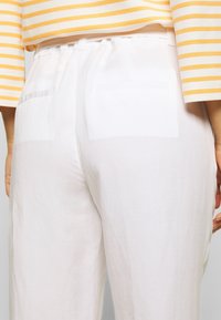 White linen pants with a loose fit, featuring a smooth texture and two back pockets. The model is wearing a striped yellow and white top.