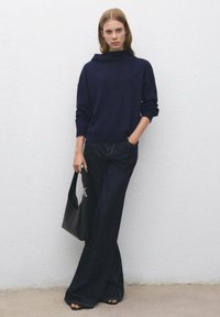 Donna che indossa un maglione blu scuro e jeans a gamba larga, che tiene una borsa nera, in piedi contro un muro bianco texture.