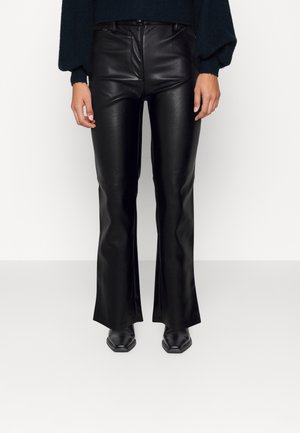 Vue de face d'une personne portant un pantalon évasé en similicuir noir, des bottes noires à bout pointu et un haut à manches longues foncé avec des manches bouffantes.