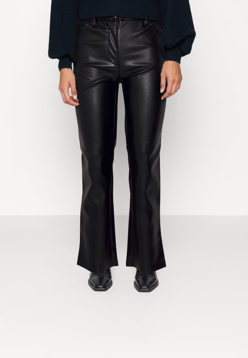 Vue de face d'une personne portant un pantalon évasé en similicuir noir, des bottes noires à bout pointu et un haut à manches longues foncé avec des manches bouffantes.