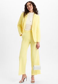 Gelbes Blazer und passende weit geschnittene Hose aus leichtem Stoff. Weißes Oberteil mit Verzierungen. Silberne Handtasche und High Heels.