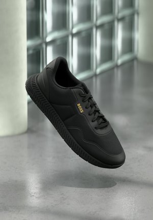 Zapatilla negra BOSS con suela texturizada y diseño de cordones, flotando sobre un suelo gris reflectante cerca de una pared de bloques de vidrio.