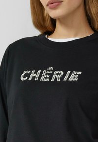Czarna bluza z perłowymi akcentami tworzącymi napis "CHÉRIE" w wyrazistym, zabawnym wzorze na przodzie. Miękki materiał, okrągły dekolt.