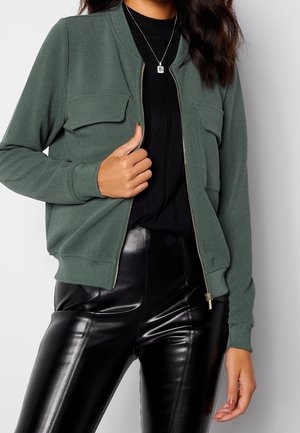 Mujer lleva puesta una chaqueta verde oscuro con cremallera sobre un top negro y pantalones de cuero negro brillantes, sujetando la cremallera de la chaqueta con una mano.