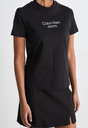 Femme portant un t-shirt noir à col rond avec le texte « Calvin Klein Jeans » imprimé en blanc, associé à une jupe noire fendue à l'avant.