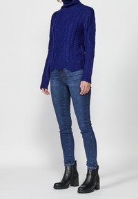 Pull bleu en maille avec un col montant côtelé, présentant des motifs torsadés et un ourlet festonné. Associé à un jean en denim foncé et des bottines noires.