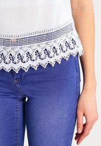 Blusa bianca con rifiniture in pizzo decorativo sulla parte inferiore, abbinata a jeans in denim blu. Il pizzo presenta un bordo ondulato e motivi intricati.